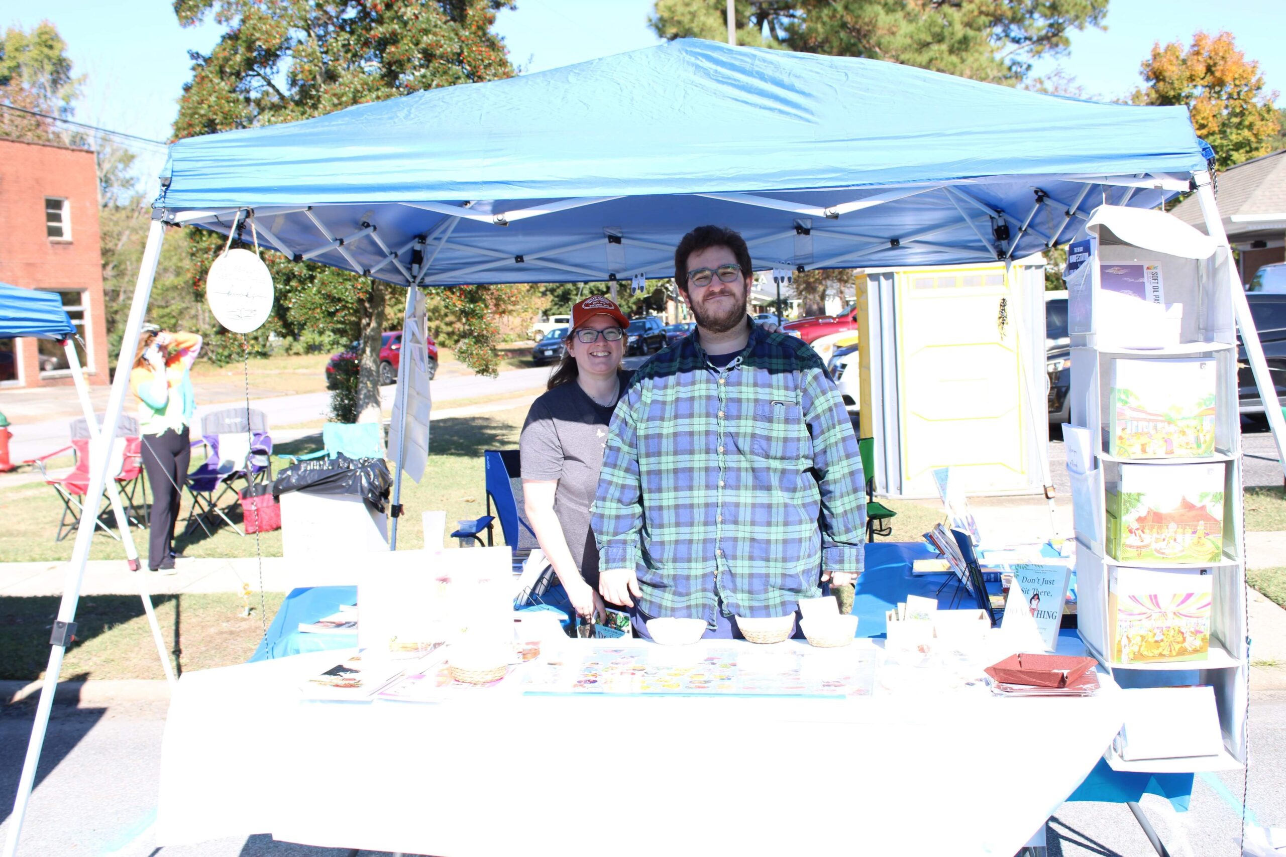 James and Elisabeth at the Rebuild Better Life Books Stand under a blue canopy behind a table of stickers and books